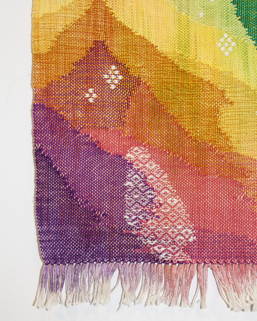 A close up of the bottom left corner of the wall hanging shows white woven diamonds overlaid on top of a background in purples and oranges.