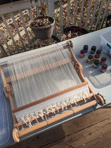 an image of a loom warped with white yarn and a selection of paints on the table next to it.