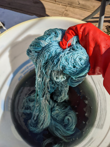 a gloved hand removing teal yarn from a bucket of murkey liquid.