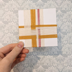 A 4 inch square card wrapped with overlapping strands of yarn in cream, yellow and pink.