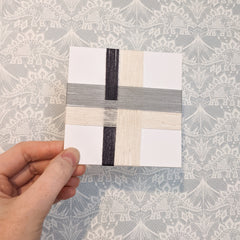 A 4 inch square card wrapped with overlapping strands of yarn in cream, pale blue gray and dark charcoal gray.