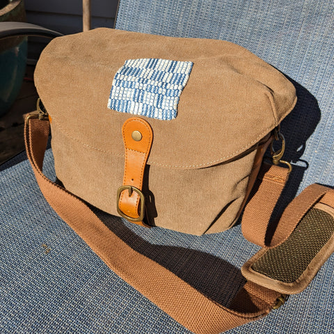 A brown canvas camera bag sits in a chair in the sun. Front and center, right where a logo would normally be placed, a small square patch has been sewn on. The patch features blue and cream squares, not unlike a quilt.