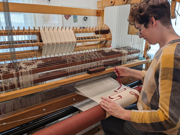 An image of a white person with short red hair sitting at a loom, yarn in hand.