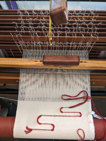 A top down image of a table runner mid-weave on a loom. The runner has a natural cotton background color, and half of a red squiggle has been inlaid in the cloth with the tail of the red yarn hanging out.