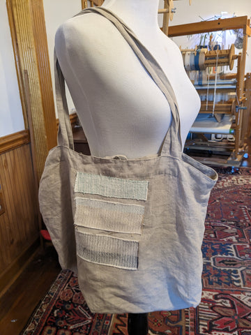 A beige canvas tote bag has been decorated with three pieces of handwoven cloth sewn to the outside. The patches are in pale shades of grey, salmon and blue and layered like bricks.