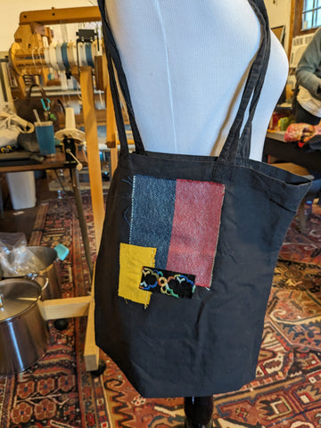A black tote bag sits on the shoulder of a mannequin. It has three bright patches that have been sew on the surface, overlapping. A small black square with rainbown embroidery over top a bright yellow square over top the largest rectangle that is split blue and red.