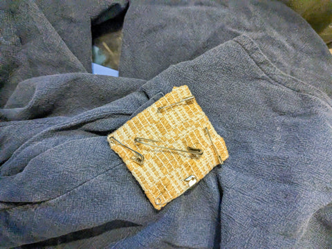 A close up photo of a yellow patch safety pinned to blue cloth, covering a hole. There are five safety pins on the small yellow patch, one at each corner and an extra large pin in the middle.