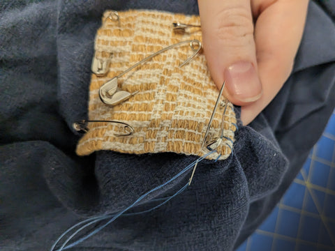 The same yellow patch on blue fabric, now there is a needle stiching along one edge of the patch attaching it to the fabric.