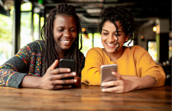 Two women sitting at a table showing each other what is on their phone screens.