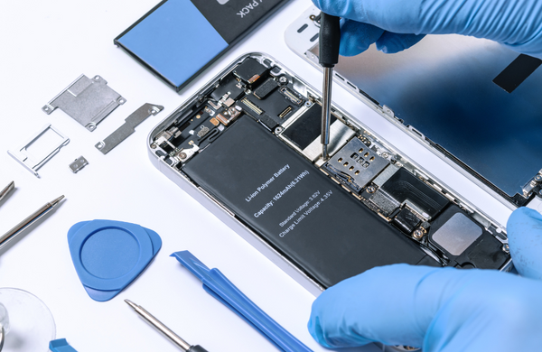 Two hands wearing blue gloves refurbishing a smartphone. One hand is holding a small screw driver tool and the other is holding the smartphone, which does not have a screen and the inside technology (the battery etc.) is visible. There are other refurbishing tools and parts laid out on the bench around the phone.