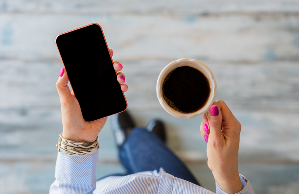 A woman with pink fingernails holding a smartphone and mug of coffee.