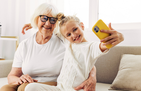 A grandmother posing next to her granddaughter while she takes a selfie on a phone with a yellow case. 