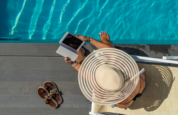 A women dipping her toes into a pool holding a smartphone and wearing a straw hat.