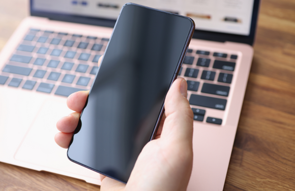 A hand holding a Samsung phone with a black display screen in front of an open rose gold laptop.