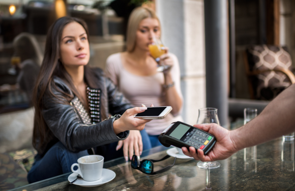 Two women sitting at a table. One of the women is in the background sipping on an orange juice and the other woman is holding her phone up to an Eftpos machine which is held out by the server. There is two empty coffee cups on the table and an empty glass. 