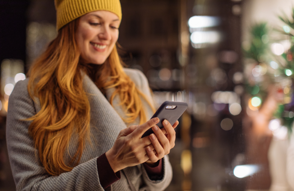 A woman wearing a yellow beanie and grey coat looking at the screen of a Samsung phone holding it in both hands.