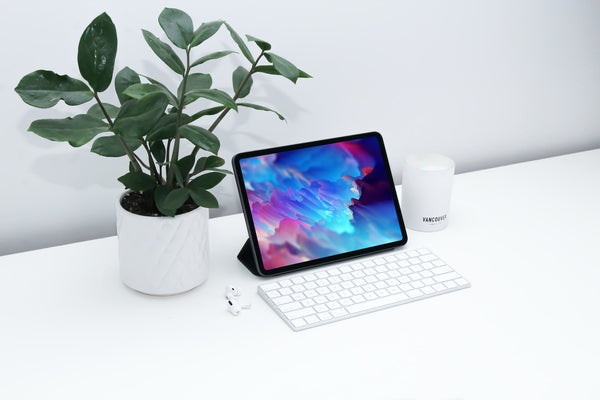 Black iPad standing up next to airpods, a white candle and a white keyboard