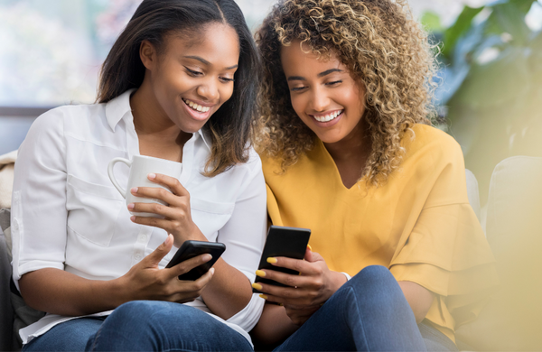 Two woman looking down at their phone and smiling. One is also holding a coffee cup wearing a white t-shirt and the other is wearing a yellow shirt..