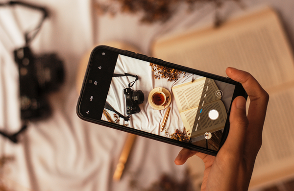 A hand holding a Samsung smartphone with the camera app open, focusing on a traditional camera, tea and book.