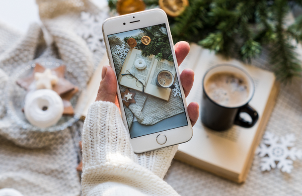 A white iPhone being held in a hand covered by a white knitted jumper sleeve with a coffee and book in the background.