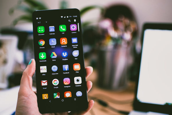 A hand holding an Android phone with the Apps displayed on the screen. There is a home office out of focus in the background.