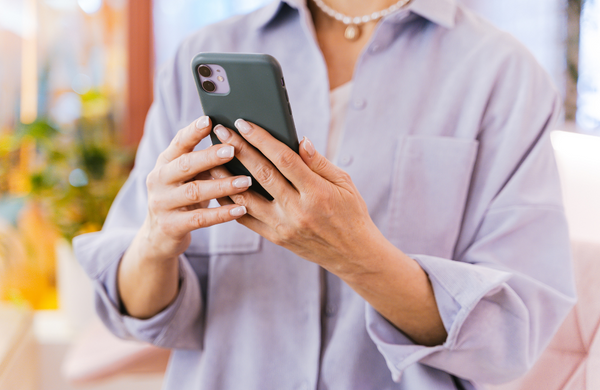 A middle aged woman holding an iPhone with a forest green case in two hands. Her face is not visible in the image but she is wearing a dusty lilac button up and strong of pearls around her neck. 