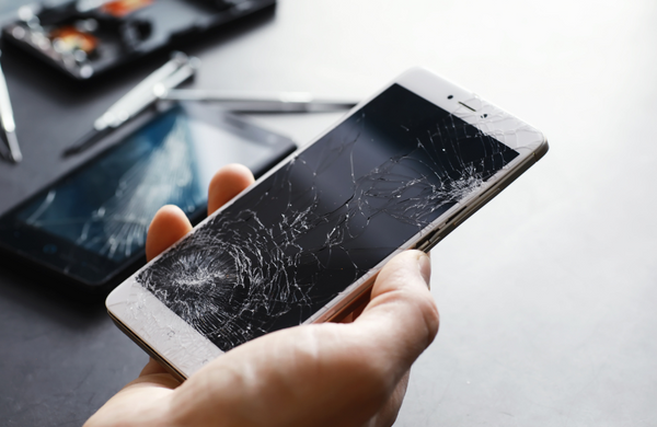 A hand holding a smartphone with a cracked screen. There are repair tools and other phones with cracked screens in the background.