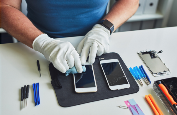 A person wearing white cloves cleaning the dust off of a new screen with an old cracked screen next to it. There is also various phone refurbishing tools laid out on the bench. 