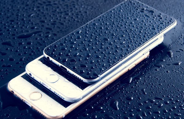 Three different models of iPhone stacked on top of each other on top of a black surface. There are water droplets covering the phones and black surface.