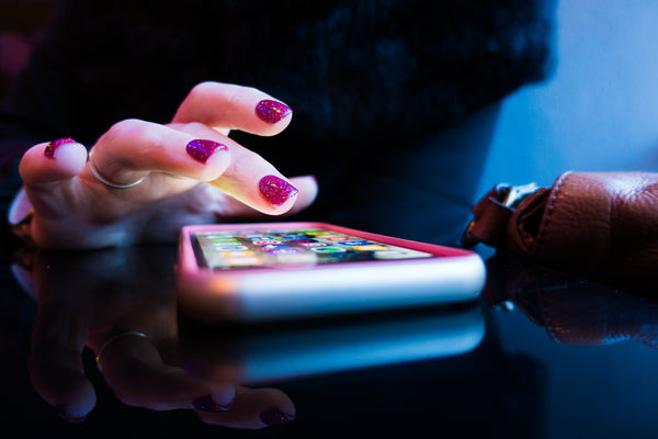 A phone on a table with a hand with glittery pink painted nails hovering over the top of the screen.