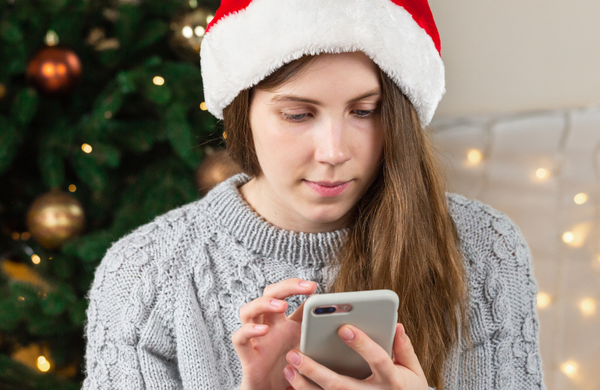 A woman wearing a Santa hat looking down at her phone in front of a Christmas tree.