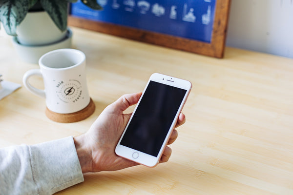 A hand holding an iPhone with a blank screen over a desk next to a coffee mug.