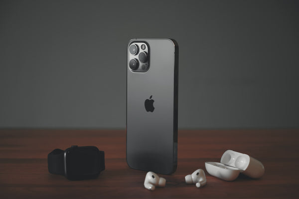 A black iPhone standing upright on a wooden desk next to Air Pods and an Apple watch.