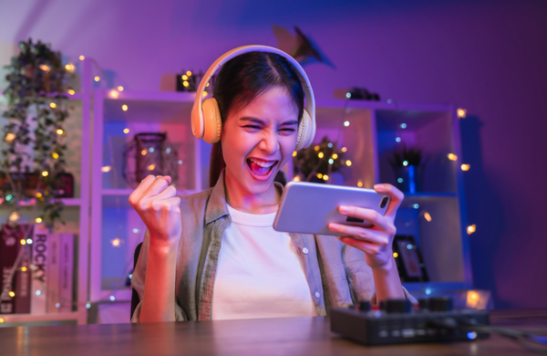 A young teenage girl with headphones on looking at a smartphone and clenching her fist in excitement.