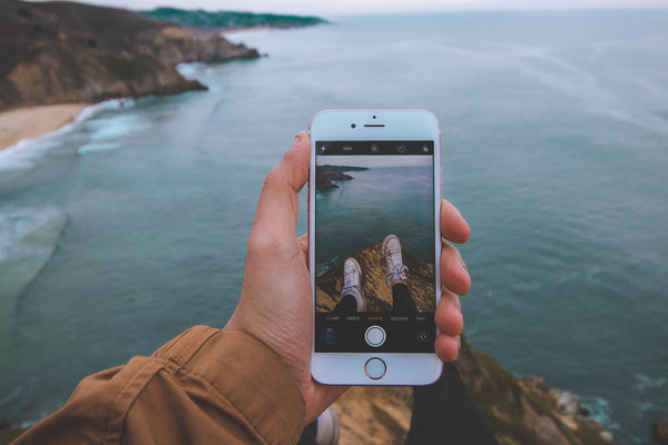 An iPhone screen with the camera open pointing at a person sitting on a cliff edge above the ocean.