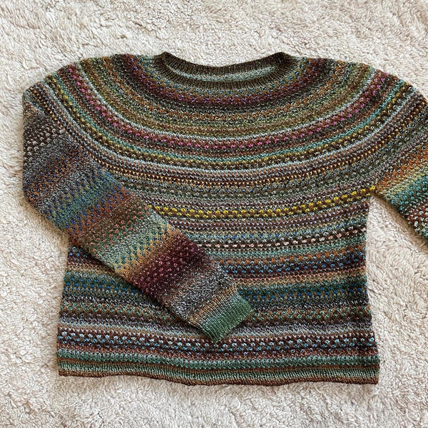 Ein Rundpassenpullover liegt auf dem Boden, es sind nur Teile davon zu sehen. das Pattern ist "Shifty" von Andrea Mowry, gestrickt mit Crazy Zauberbällen von Schoppel in warmen Braun- und Grüntönen.