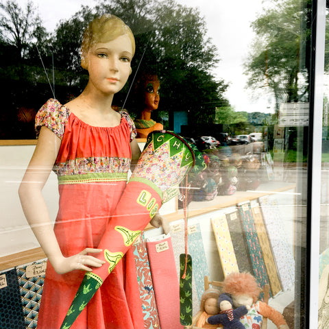 Eine blonde Mädchenschaufensterpuppe hat ein selbst genähtes orangenes Kleid an und hält eine selbst genähte Dinosaurierschultüte in den Armen. Die Schultüte ist aus den gleichen Stoffen genäht wie das Kleid. Spiegelungen im Bild zeigen, dass die Schaufensterpuppe von außen durch eine Glasscheibe fotografiert wurde.