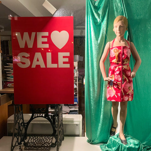 Aufnahme durch eine Geschäftsschaufensterscheibe mit einem roten "we love Sale Plakat links und einer blonden Schaufensterpuppe im roten Kleid rechts.