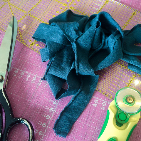 Blauer schmaler Stoffstreifen in einem kleinen Haufen liegend. Daneben eine Metallstoffschere und ein grüngelber Rollschneider.