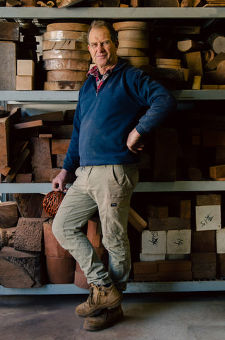 A tall man turned towards the camera dressed in a blue jumber with khaki work pants and heavy, desert boots stands in front of three shelves stacked with various sized & shaped wooden objects.