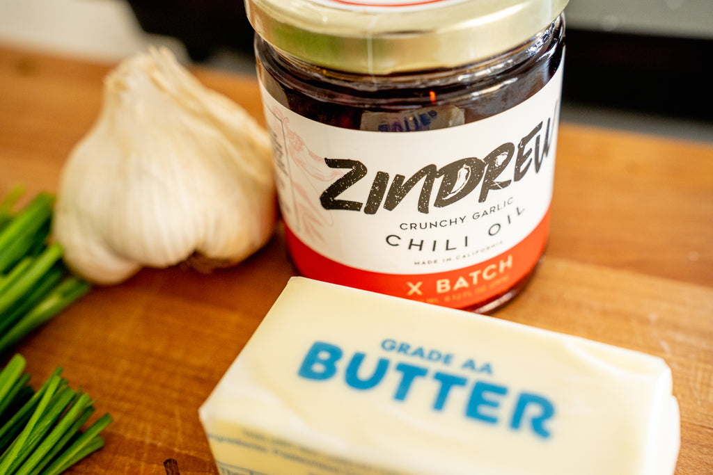 Zindrew's Garlic Chili Oil on a wooden cutting board by a head of garlic and a stick of butter