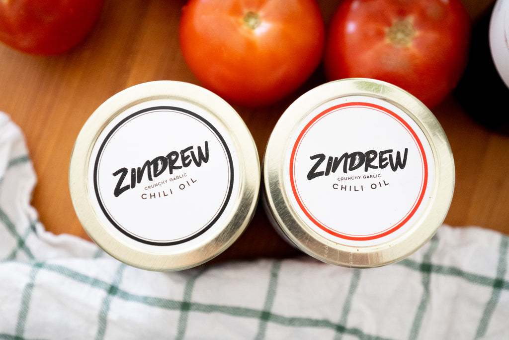 zindrew garlic chili oil jars on a cutting board with a kitchen towel and tomatoes