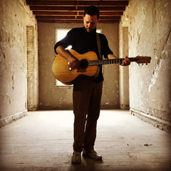 Kyle Rasche standing in a empty room with his guitar. 