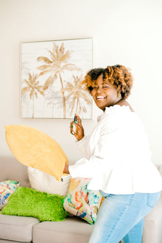 Black woman turned to the side smiling with a white shirt and jeans while holding a yellow pillow and spritzing room and body spray on the yellow pillow.