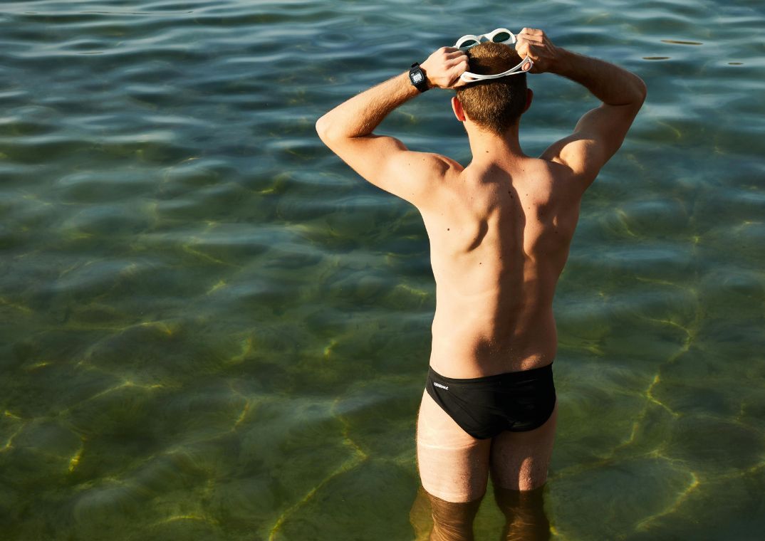man standing in water wearing swimming trunks with back to camera