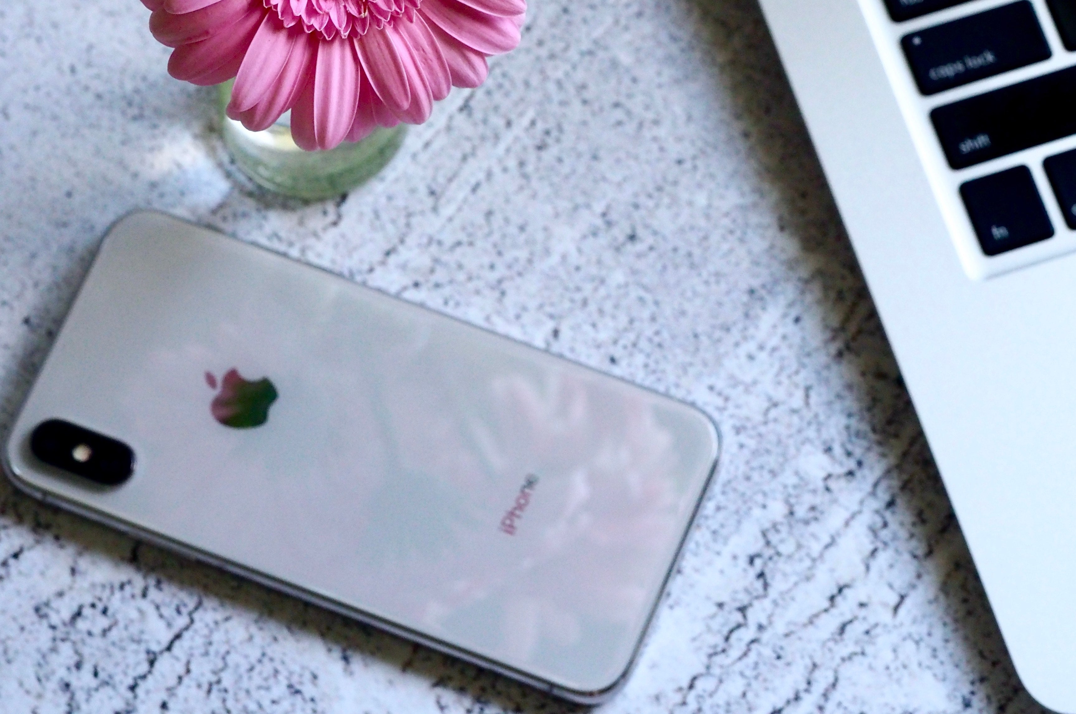 A silver iphone X on a wooden table next to pink flowers and a macbook.