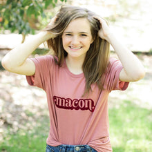Load image into Gallery viewer, Photo of girl smiling wearing a pink tee with &quot;macon&quot; printed in the center with greenery in the background
