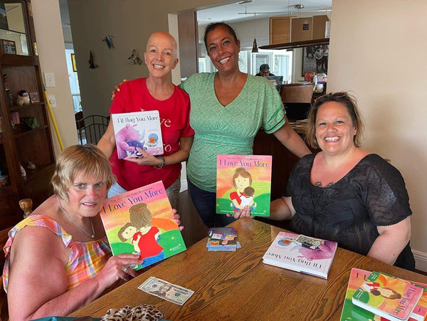 Laura Duksta smiles next to a kitchen table with three friends holding copies of her books that will be donated to a NICU.