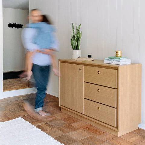 father in jeans and shirt carrying his young child who's back is to the camera, in front of a Forti Goods Eleanor lockable cabinet in oak wood.