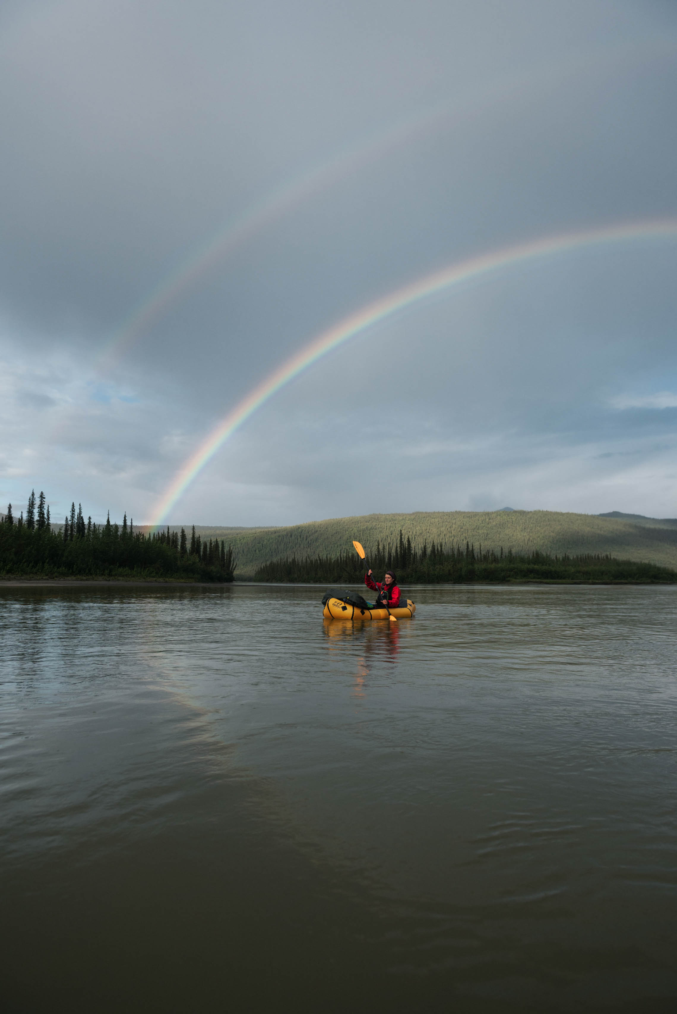 Person packrafting in an arctic lake with a double rainbow above